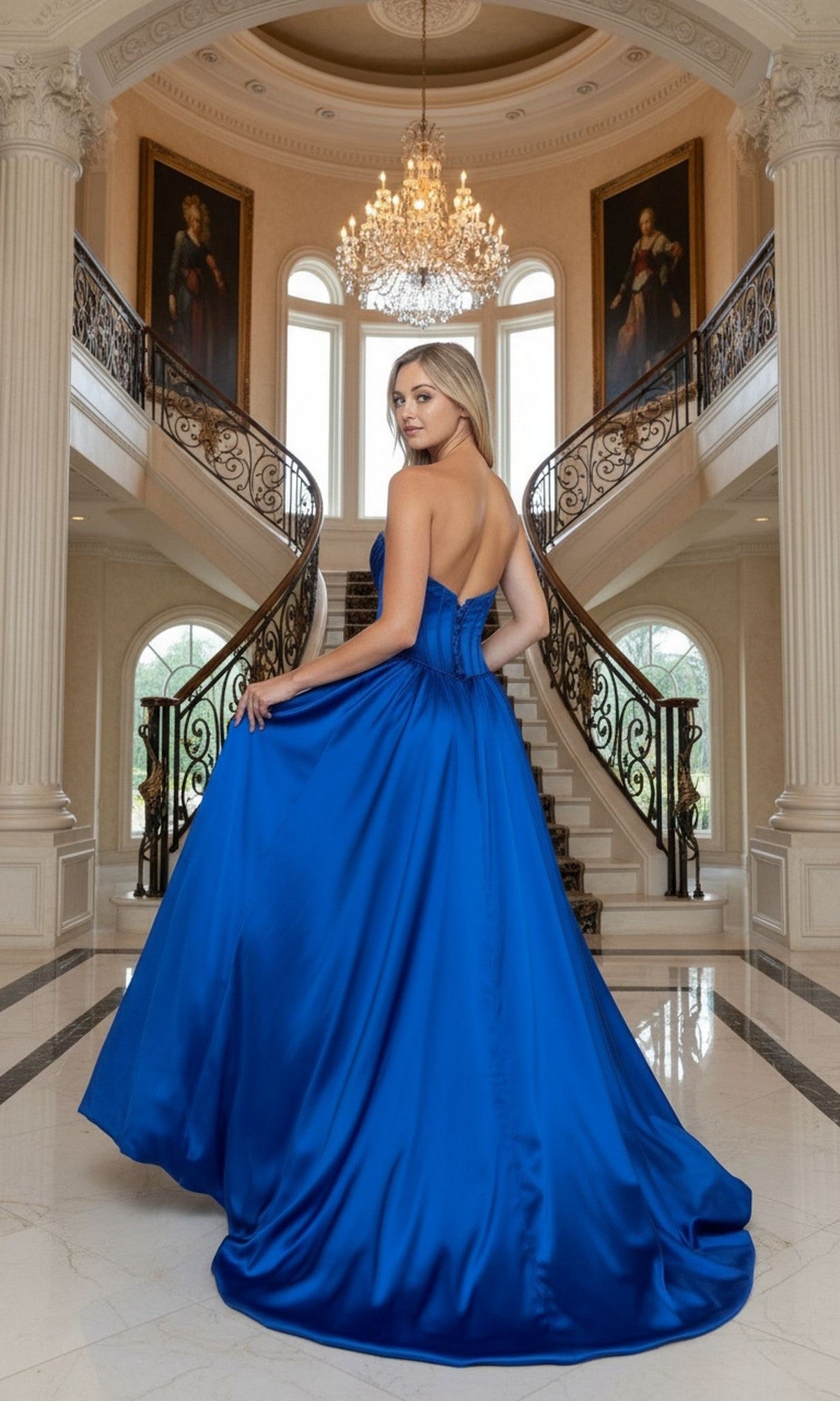 A woman in the Poly USA 25136 long prom dress, featuring a strapless royal blue corset bodice, stands before a grand staircase with ornate railings and sparkling chandeliers in an opulent setting.